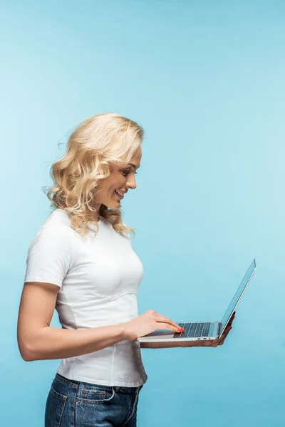 Happy curly blonde woman using laptop while standing on blue — Stock Photo