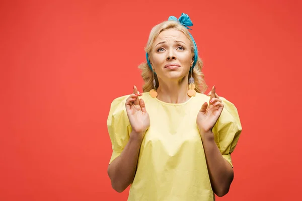Triste femme blonde en bleu bandeau debout avec les doigts croisés sur rouge — Photo de stock