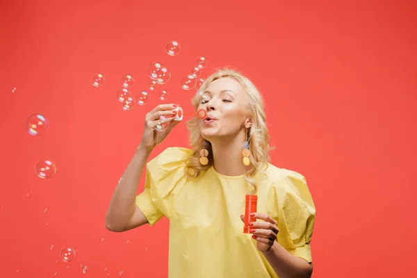 Cheerful blonde woman blowing soap bubbles on red — Stock Photo