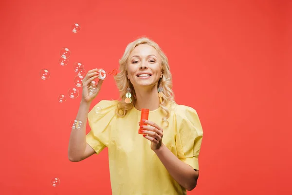 Cheerful blonde woman holding bottle near soap bubbles and smiling on red — Stock Photo