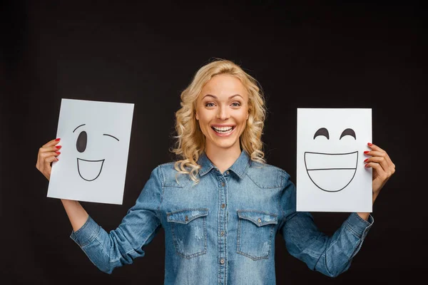 Cheerful blonde woman holding papers with happy faces isolated on black — Stock Photo