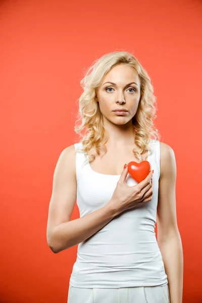 Attractive blonde woman holding heart while looking at camera on red — Stock Photo