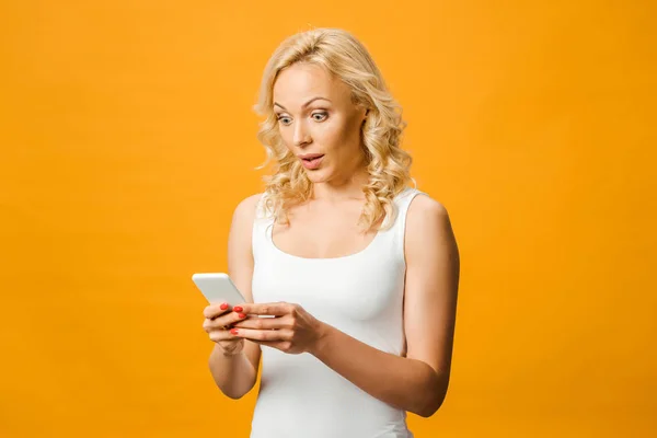 Здивована блондинка дивиться на смартфон ізольовано на помаранчевому — стокове фото