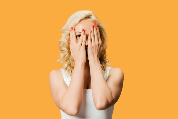 Кучерява блондинка прикриває обличчя і дивиться на камеру ізольовано на помаранчевому — стокове фото