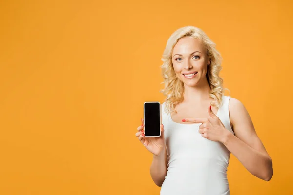 Felice donna riccia che punta con il dito verso smartphone con schermo bianco isolato su arancione — Foto stock