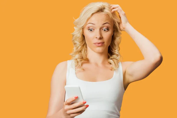 Surpris femme blonde regardant smartphone et toucher les cheveux isolés sur orange — Photo de stock