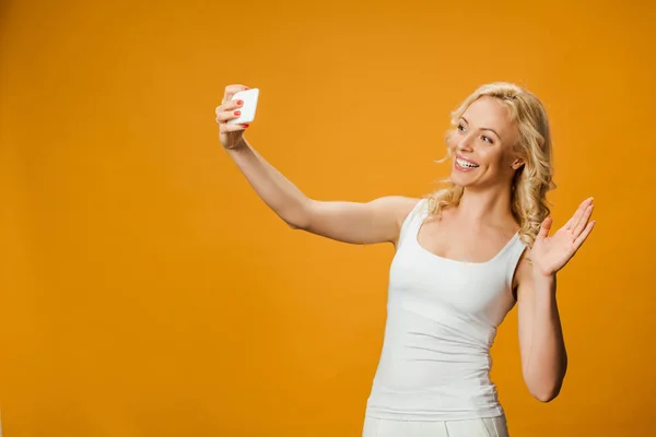 Fröhliche blonde Frau winkt mit der Hand, während sie Selfie auf orange macht — Stockfoto
