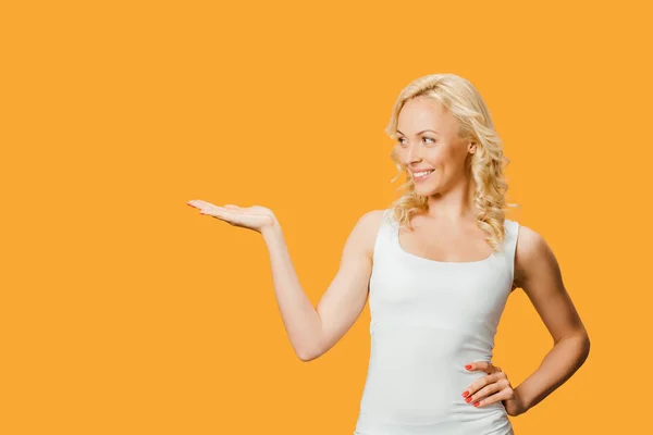 Bella donna bionda gesticolare mentre in piedi con mano sul fianco isolato su arancione — Foto stock