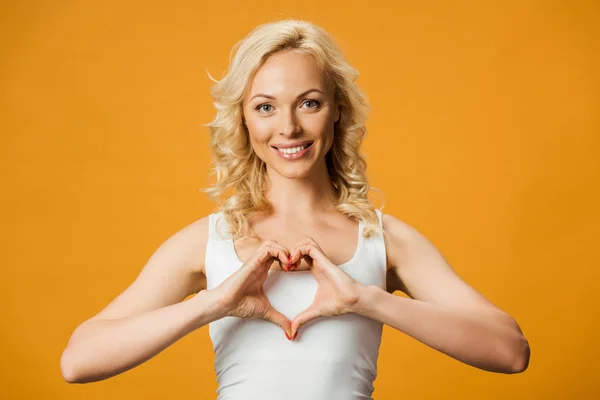 Femme blonde gaie montrant symbole en forme de coeur sur orange — Photo de stock