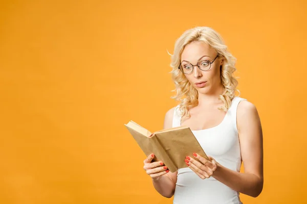 Mujer rubia sorprendida en gafas libro de lectura aislado en naranja - foto de stock