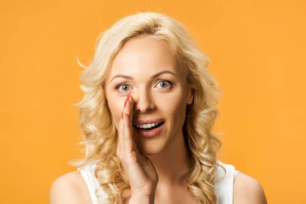 Mulher loira atraente olhando para a câmera e fofocando isolado na laranja — Fotografia de Stock