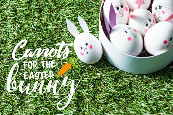 Chicken eggs with funny bunny faces and paper ears in bowl on green grass background with carrots for easter bunny lettering — Stock Photo