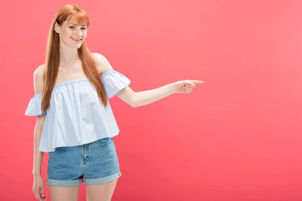 Smiling redhead woman in denim shorts pointing with finger isolated on pink — Stock Photo