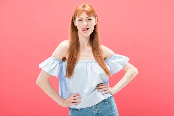 Front view of confident redhead girl standing with hands on hips and sticking out tongue isolated on pink — Stock Photo