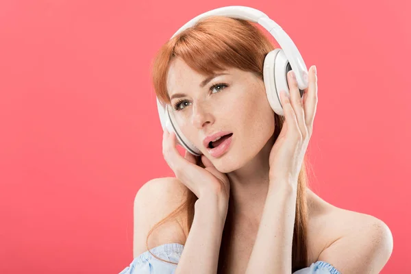 Attrayant rousse femme écouter de la musique dans les écouteurs et regarder loin isolé sur rose — Photo de stock