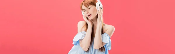 Panoramic shot of attractive redhead woman listening music in headphones with closed eyes isolated on pink — Stock Photo