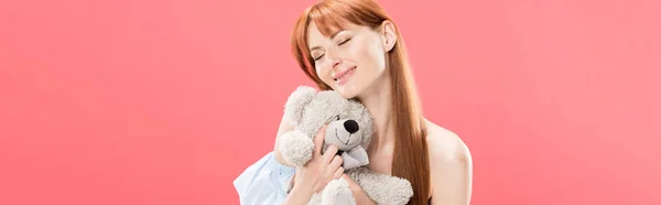 Plan panoramique de jeune femme rousse souriante tenant un ours en peluche les yeux fermés isolé sur rose — Photo de stock