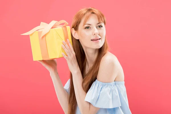 Curieuse rousse anniversaire fille tenant boîte cadeau jaune avec ruban isolé sur rose — Photo de stock