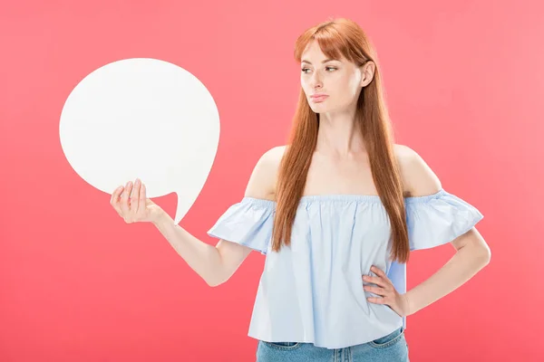 Serious attractive redhead girl standing with hand on hip and holding blank speech bubble isolated on pink — Stock Photo
