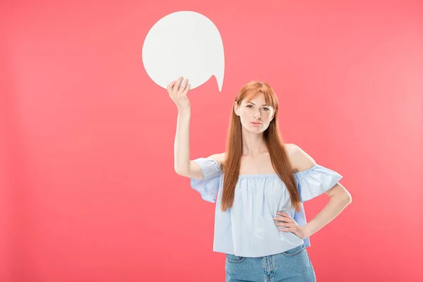 Front view of serious attractive redhead girl standing with hand on hip and holding blank speech bubble isolated on pink — Stock Photo