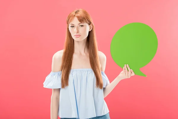 Serious attractive redhead girl holding green blank speech bubble and looking at camera isolated on pink — Stock Photo