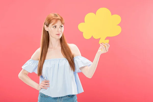 Attractive redhead young woman standing with hand on hip and holding empty thought bubble isolated on pink — Stock Photo