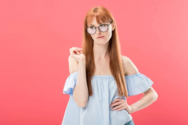 Pensive attractive redhead young woman in glasses standing with hand on hip isolated on pink — Stock Photo