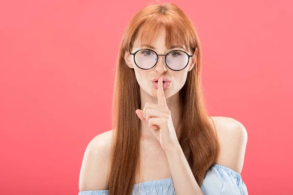 Front view of redhead girl in glasses looking at camera and showing hush sign isolated on pink — Stock Photo