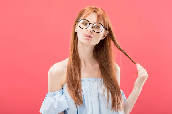 Front view of redhead girl in glasses and stylish blouse playing with hair and looking at camera isolated on pink — Stock Photo