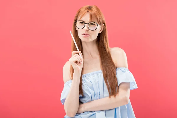 Pensive redhead young woman in glasses holding pencil isolated on pink — Stock Photo