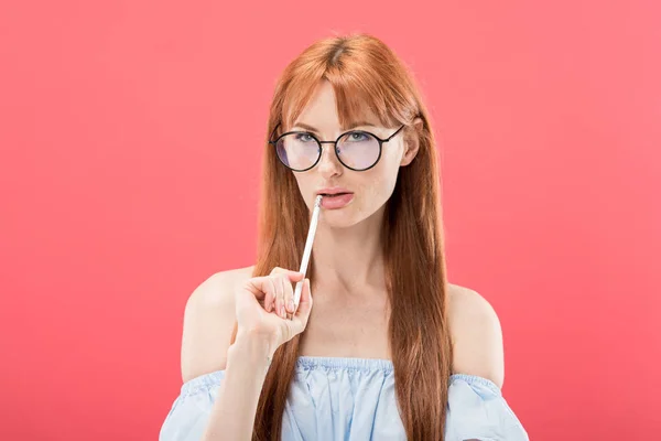 Coûteuse rousse jeune femme dans des lunettes tenant crayon isolé sur rose — Photo de stock