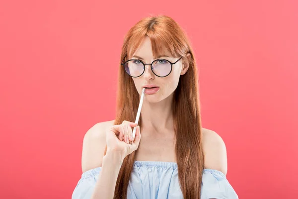 Pensativo pelirroja joven en gafas celebración lápiz aislado en rosa - foto de stock