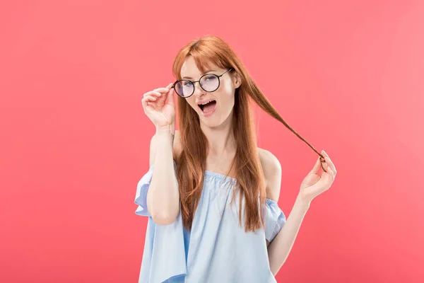 Улыбающаяся рыжая девушка в очках и стильная блузка, играющая с волосами и смотрящая на камеру, изолированную на розовом — стоковое фото