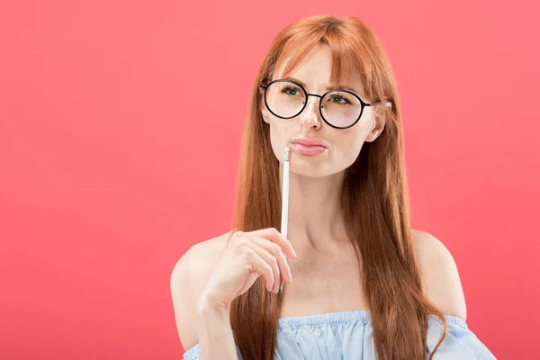 Pensativo pelirroja joven en gafas celebración lápiz aislado en rosa - foto de stock