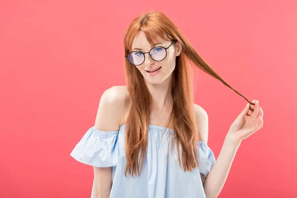 Sorridente rossa ragazza in occhiali e camicetta elegante giocare con i capelli e guardando la fotocamera isolata su rosa — Foto stock