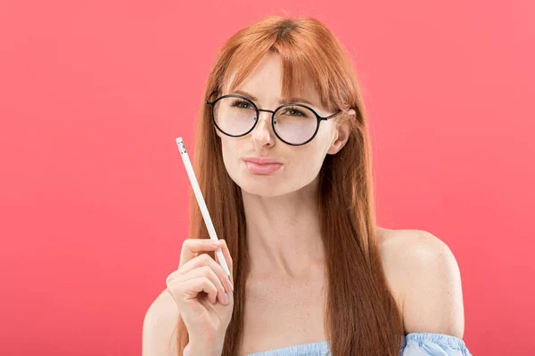 Rossa pensosa giovane donna in occhiali che tiene la matita isolata su rosa — Foto stock