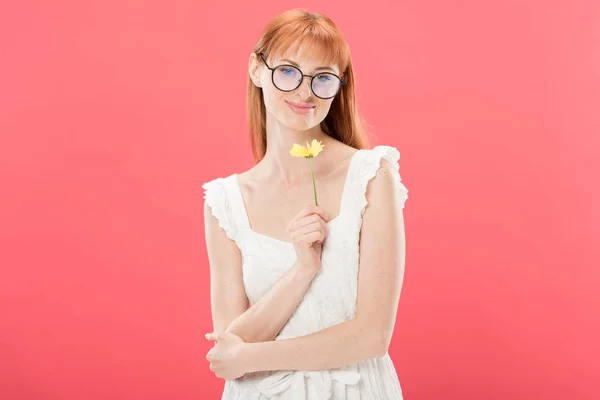 Vorderseite der schönen rothaarigen jungen Frau in weißem Kleid mit gelben Blume und Blick auf Kamera isoliert auf rosa — Stockfoto