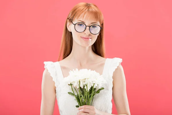 Vista frontal de chica pelirroja atractiva en gafas y vestido blanco sosteniendo flores y sonriendo aislado en rosa - foto de stock