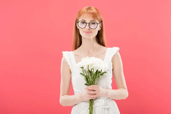 Vorderseite des attraktiven rothaarigen Mädchens mit Brille und weißem Kleid, das Blumen hält und isoliert auf rosa lächelt — Stockfoto