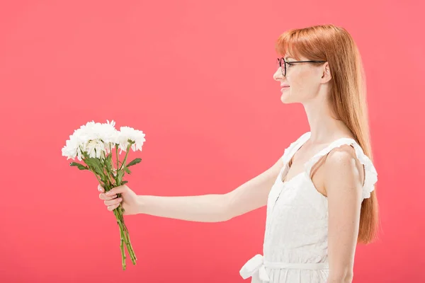 Seitenansicht des attraktiven rothaarigen Mädchens mit Brille und weißem Kleid, das Blumen hält und isoliert auf rosa lächelt — Stockfoto