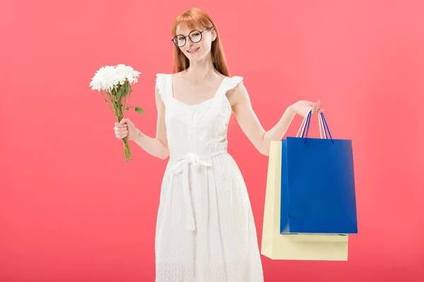 Улыбающаяся рыжая девушка в очках и белом платье держа букет цветов и торговых мешков изолированы на розовый — стоковое фото