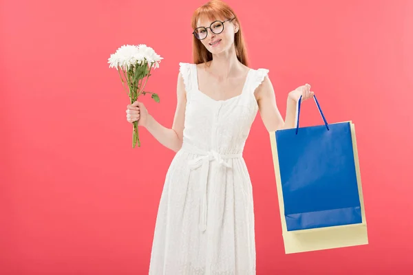 Smiling redhead girl in glasses and white dress holding bouquet of flowers and shopping bags isolated on pink — Stock Photo