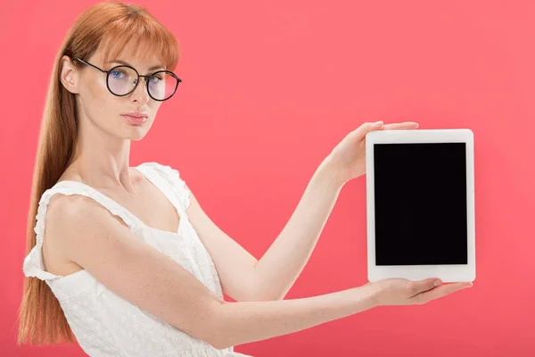 Hermosa pelirroja en gafas y vestido blanco sosteniendo tableta digital con pantalla en blanco y mirando a la cámara aislada en rosa - foto de stock