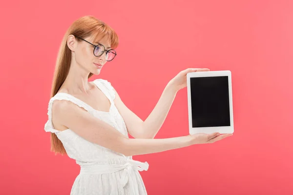 Красива руда дівчина в окулярах і біла сукня тримає цифровий планшет з порожнім екраном ізольовано на рожевому — стокове фото
