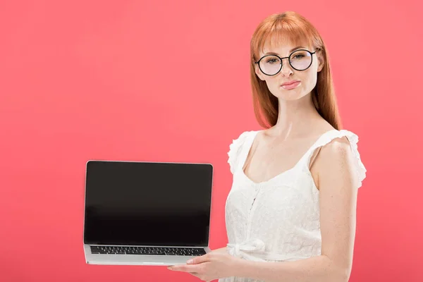 Руда молода жінка в окулярах тримає ноутбук з порожнім екраном ізольовано на рожевому — стокове фото