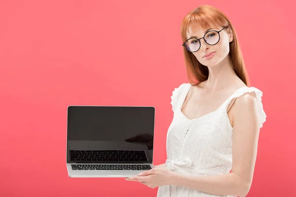 Pelirroja joven en gafas sosteniendo portátil con pantalla en blanco aislado en rosa - foto de stock