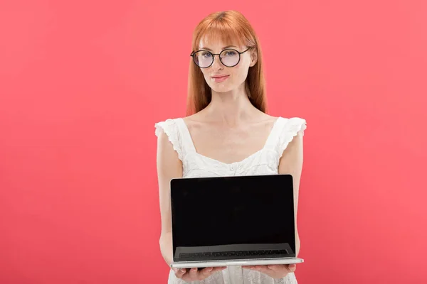 Рыжая молодая женщина в очках держит ноутбук с чистым экраном изолированы на розовый — стоковое фото