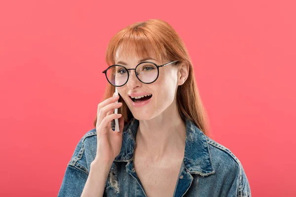 Atractiva chica pelirroja en gafas y chaqueta de mezclilla hablando en el teléfono inteligente aislado en rosa - foto de stock