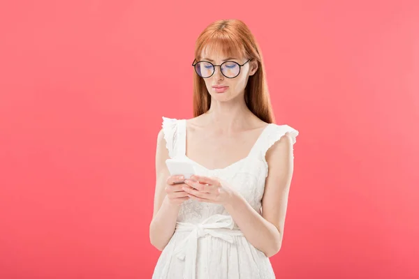Pelirroja en gafas y vestido blanco usando smartphone aislado en rosa - foto de stock