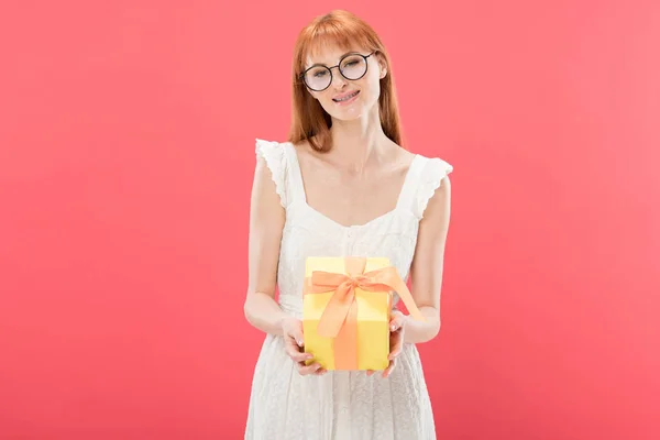 Вид спереди улыбающейся рыжеволосой девушки в очках и белом платье, которая держит подарок на день рождения и смотрит в камеру, изолированную от розового — стоковое фото